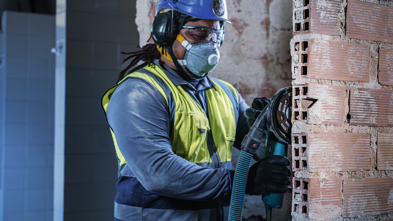 A person wearing safety equipment cuts a brick wall with a power tool.