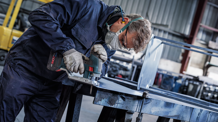 A person wearing safety equipment uses a power tool to cut sheet metal on a workbench.