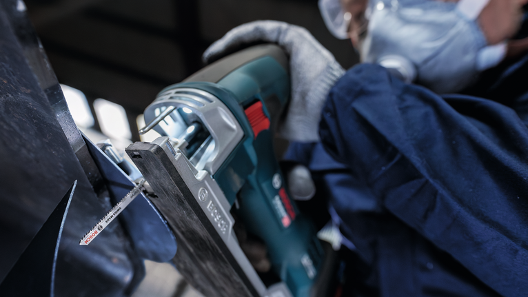 Person wearing safety equipment cuts metal with a reciprocating saw.
