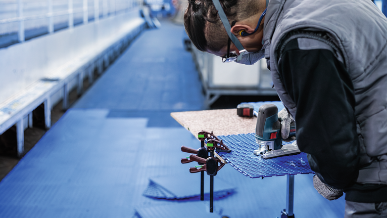 Person wearing safety equipment uses a jigsaw to cut a blue sheet clamped to a table.