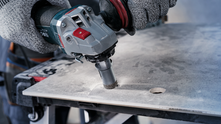 Person wearing safety equipment drills a circular hole in a tile slab.