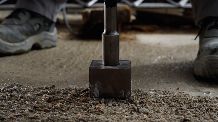 A person wearing safety equipment uses a demolition tool to break concrete.