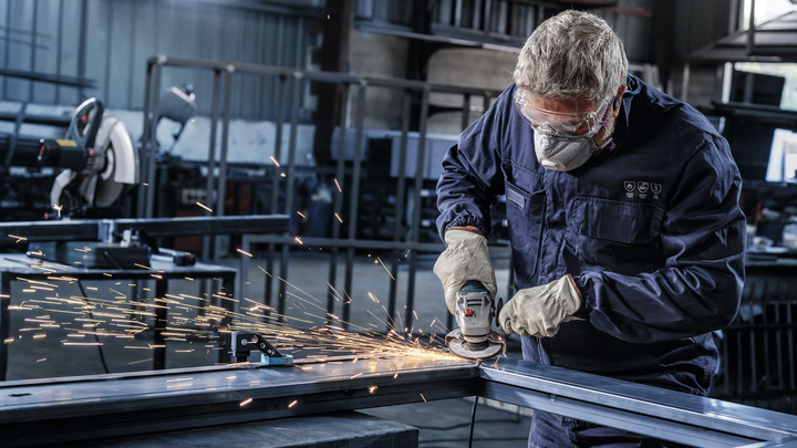 A person wearing safety equipment grinds a metal beam in a workshop, producing sparks.