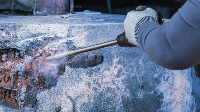 A worker wearing safety equipment chips concrete from a brick wall using a demolition tool.