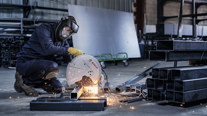A worker wearing safety equipment cuts steel beams with a large circular saw, sparks flying.