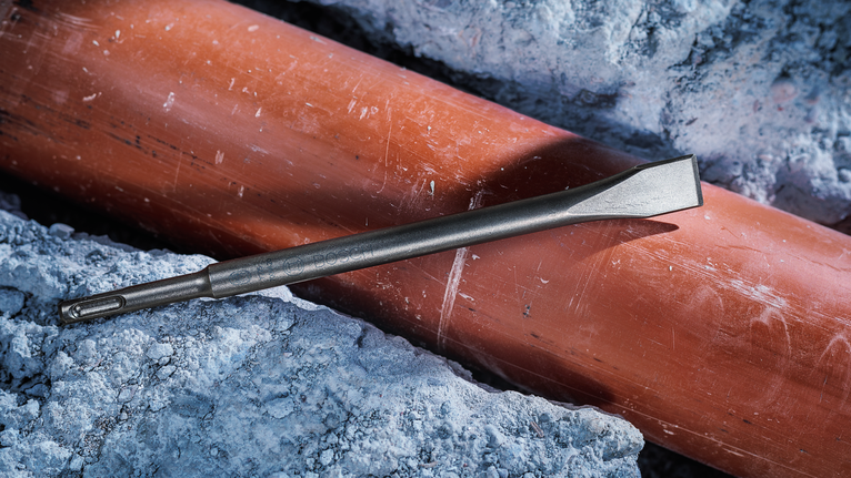 A chisel lies on a pipe surrounded by broken concrete.