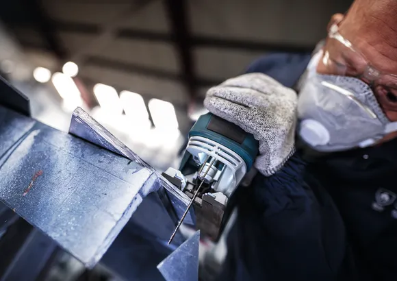 Person wearing safety equipment uses a jigsaw to cut a metal sheet.