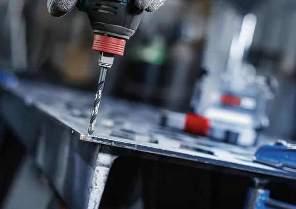 Person wearing safety equipment drills a hole in a metal sheet.