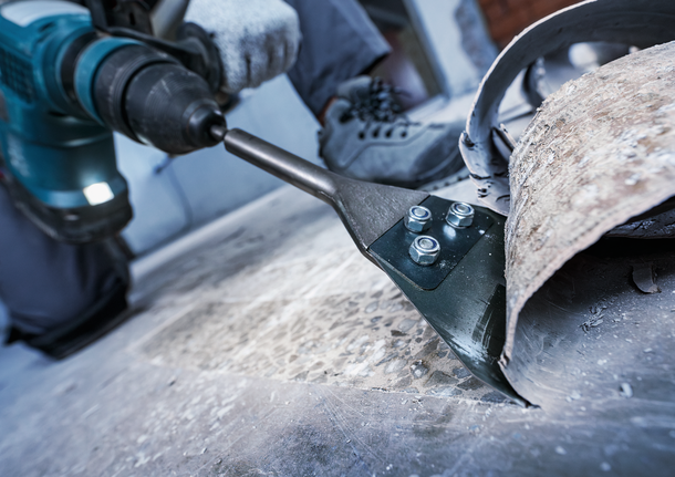 A person wearing safety equipment removes flooring material with a power chisel.