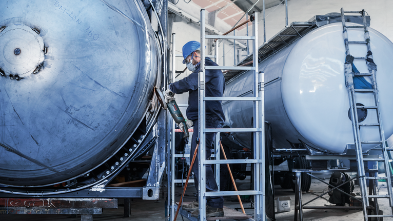A person wearing safety equipment grinds a large metal tank with a power tool.