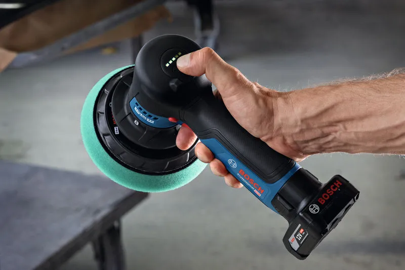 A person holds a cordless polisher with a green foam pad near a workbench.
