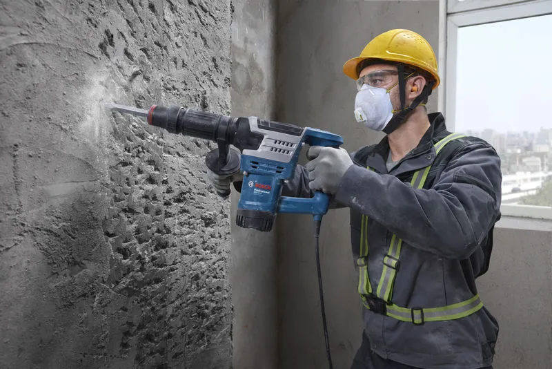 A person wearing safety equipment uses a demolition hammer to break a concrete wall.
