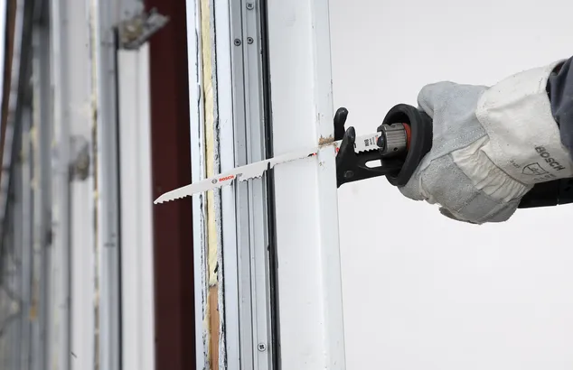 A worker wearing safety equipment uses a reciprocating saw to cut a window frame.