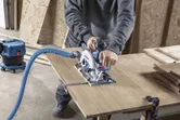 A person guides a cordless circular saw with dust extraction while cutting a wooden board.