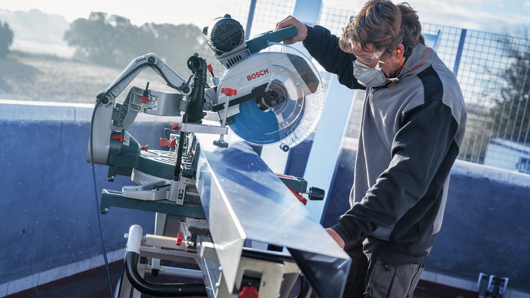 Person wearing safety equipment cuts long metal beam with a miter saw.
