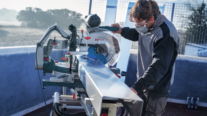 A person wearing safety equipment operates a circular saw to cut a long metal beam.