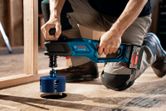 A person wearing safety equipment drills a large hole in plywood with a cordless angle drill.
