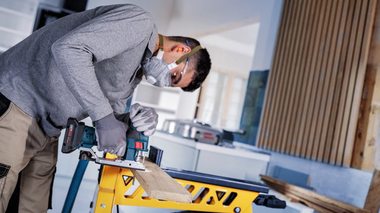 A person wearing safety equipment cuts wood with a power jigsaw on a workbench.