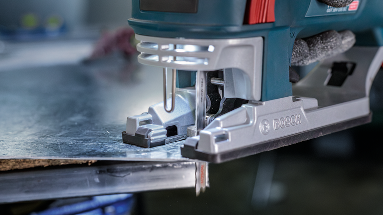 A person wearing safety equipment uses a jigsaw to cut a sheet of metal.