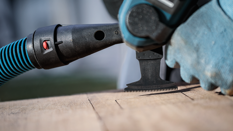 Person wearing safety equipment uses a multi-tool with vacuum attachment to cut wood.