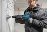 A person wearing safety equipment drills a large hole in a concrete wall using a power tool.