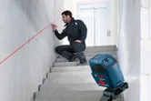 Una persona con equipo de seguridad marca una pared siguiendo la línea trazada por un nivel láser en una escalera.