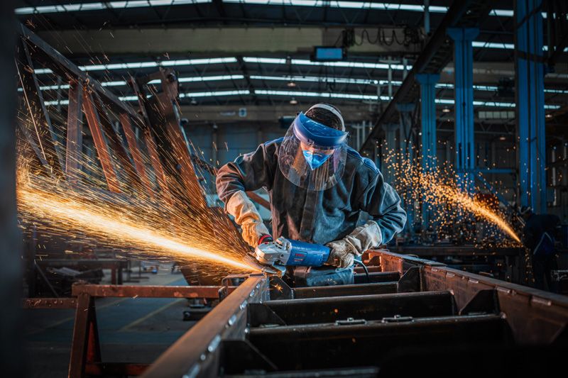 Una persona que usa equipo de seguridad muele metal con una amoladora angular, creando chispas.