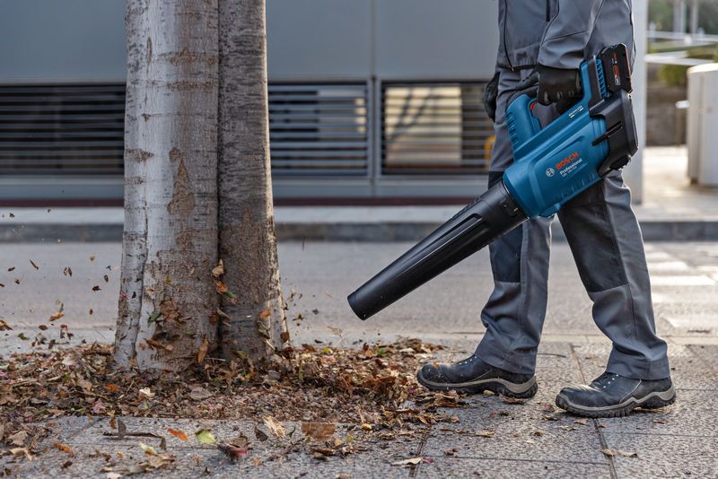 Una persona que lleva equipo de seguridad utiliza un soplador de hojas inalámbrico para mover las hojas cerca de un árbol.