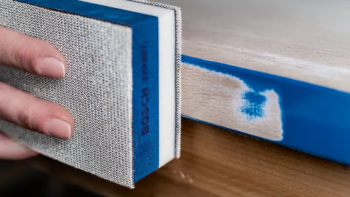 Persona lijando un borde de madera pintado con un bloque de lijado.