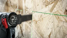 Una persona que usa equipo de seguridad utiliza una sierra alternativa para cortar madera a lo largo de una línea guía verde.