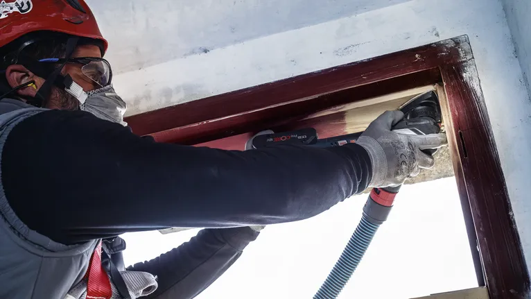 Persona que usa equipo de seguridad lijando un marco de ventana de madera con una herramienta eléctrica.