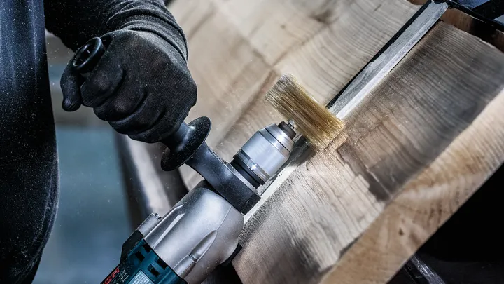 Una persona que usa equipo de seguridad utiliza una herramienta eléctrica con un cepillo de alambre para limpiar la madera.