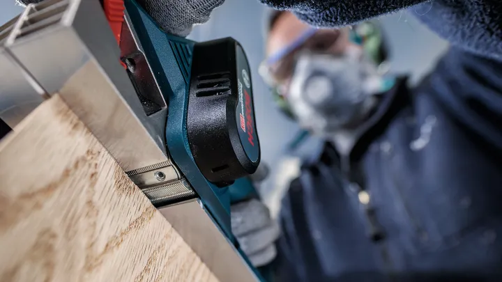 Una persona que usa equipo de seguridad utiliza una cepilladora eléctrica sobre una tabla de madera.