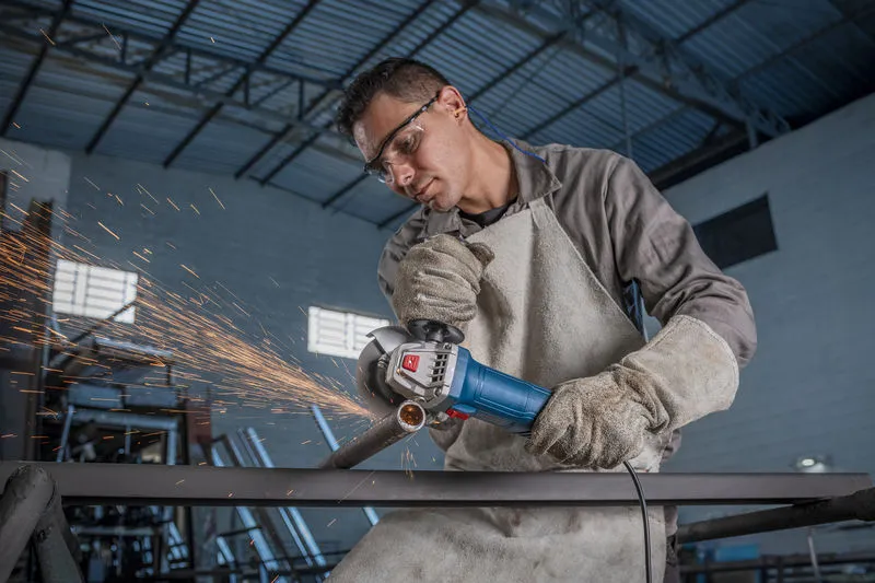 Una persona que usa equipo de seguridad muele metal con una amoladora angular, creando chispas.