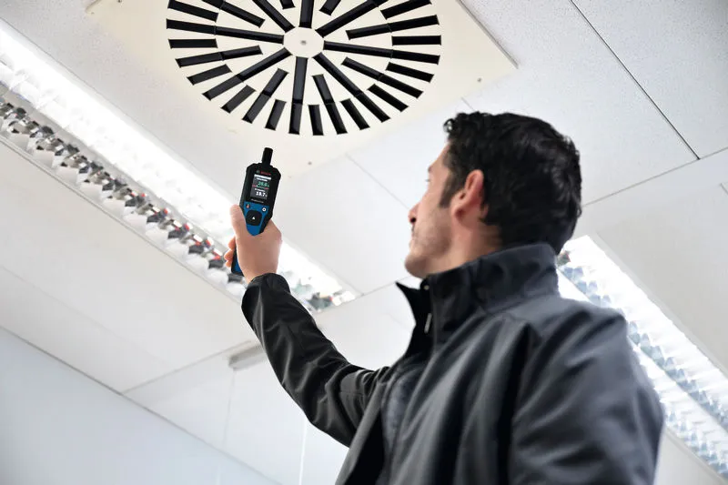Una persona sostiene un termohigrómetro cerca de una rejilla de ventilación del techo en una habitación muy iluminada.