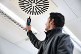 Una persona sostiene un termohigrómetro cerca de una rejilla de ventilación del techo en una habitación muy iluminada.