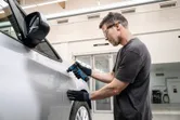 Una persona que usa equipo de seguridad lija la puerta de un automóvil utilizando una lijadora orbital.