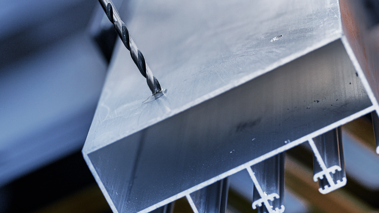 Una broca metálica que hace un agujero en un perfil rectangular de aluminio.