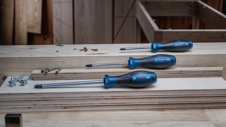 Tres destornilladores con mango azul yacen sobre tablones de madera apilados junto a varios tornillos.