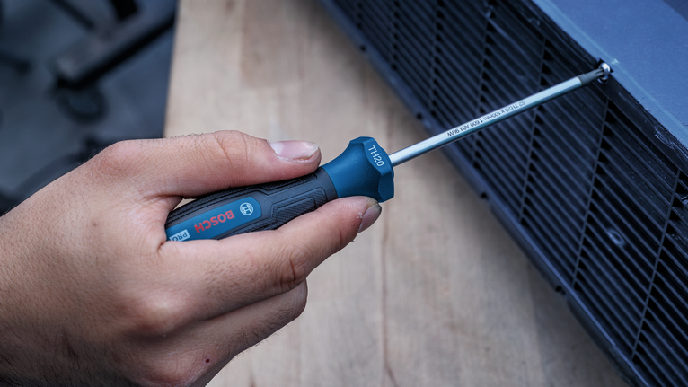 Persona que utiliza un destornillador de mango azul para apretar un tornillo en una rejilla de ventilación metálica.