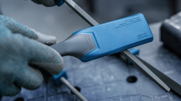 Una persona que lleva equipo de seguridad lima una varilla de metal con una herramienta de mango azul.