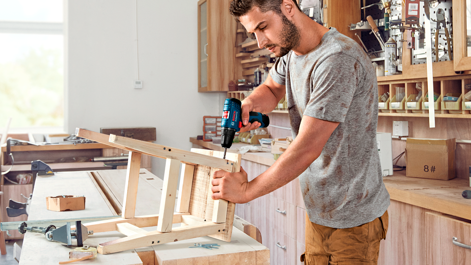Hombre usando un taladro Bosch en un taller de carpintería con muebles de madera.