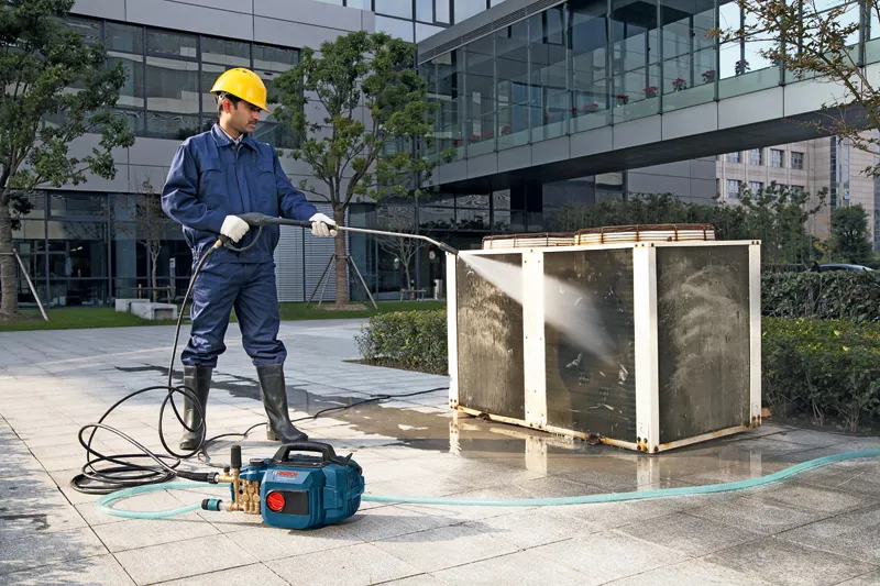 A person wearing safety equipment uses a high-pressure washer to clean an outdoor unit.