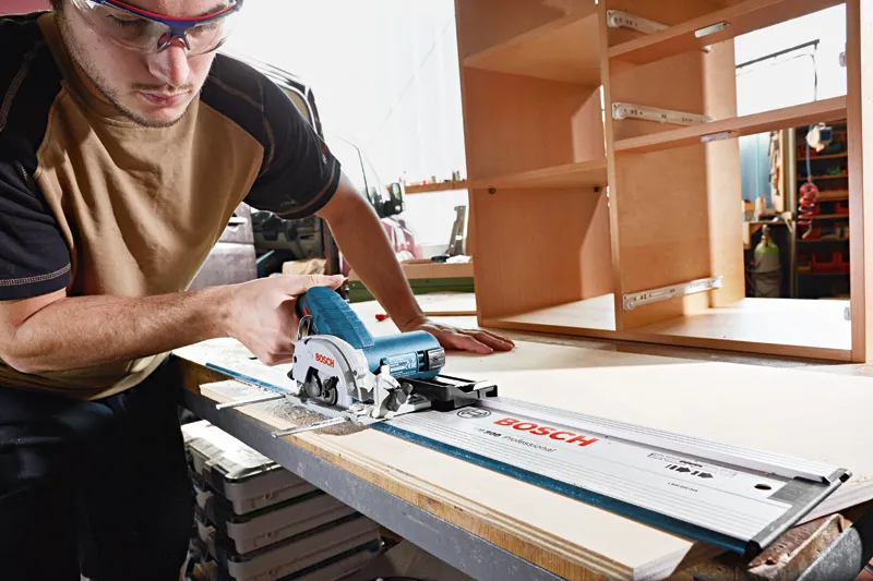 A person wearing safety equipment uses a cordless circular saw to cut wood in a workshop.