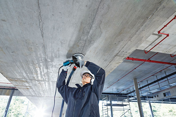A person wearing safety equipment grinds a concrete ceiling with a power tool.