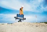 A person wearing safety equipment carries a case and tripod across a construction site.
