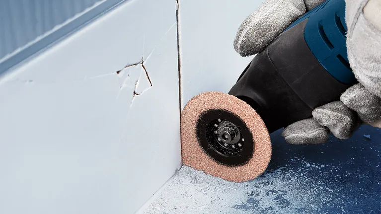 Person wearing safety equipment uses a rotary tool to grind near a wall corner.