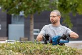 A person wearing safety equipment trims bushes using a cordless hedge trimmer.