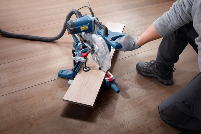 A person wearing safety equipment cuts laminate flooring with a miter saw.
