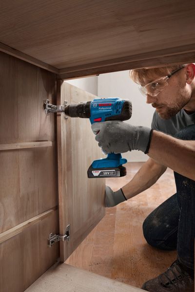 A person wearing safety equipment installs hinges on a wooden cabinet with a cordless drill.